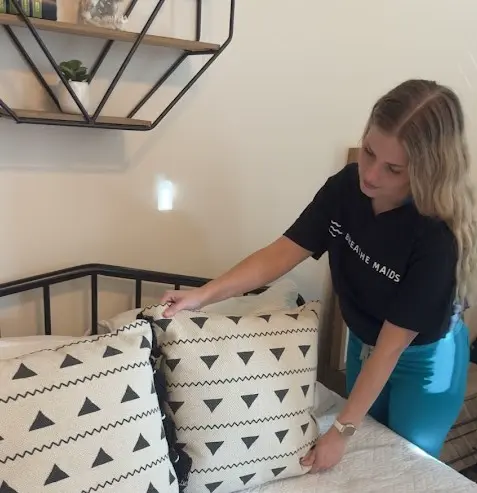 A woman arranging cushions on a bed in a well-lit bedroom, dressed in a Breathe Maids uniform, demonstrating care in room tidying