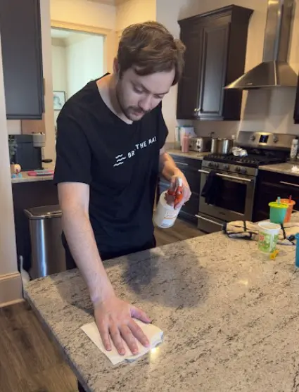 A man wiping a kitchen counter with a spray cleaner and cloth, dressed in a black Breathe Maids t-shirt, focusing on cleaning granite surfaces.