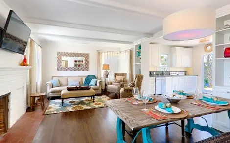 Espacioso salón comedor con piso de madera, mesa de comedor rústica puesta para seis, sofás cómodos alrededor de una chimenea blanca, y decoraciones de estilo moderno-coastal