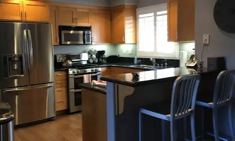 Cocina moderna con electrodomésticos de acero inoxidable, gabinetes de madera clara, y una barra con sillas altas azules, iluminada naturalmente a través de una ventana grande